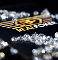 A collection of sparkling, clear gemstones surrounds a dark surface featuring the prominent logo and text 'Real Poker.' The logo design includes gold and white elements set against a black background.