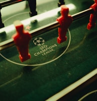 A foosball table is shown with red and black player figures positioned on the rods. The surface features the UEFA Champions League logo, indicating a themed table design.