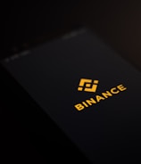 A smartphone screen displaying the Binance logo with yellow text and an icon in the center against a dark background.