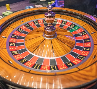 A roulette wheel with alternating red and black numbered pockets on a wooden platform. The wheel contains a small white ball resting in one of the pockets, indicating gameplay. Surrounding the wheel are stacks of colorful poker chips, suggesting a casino environment.