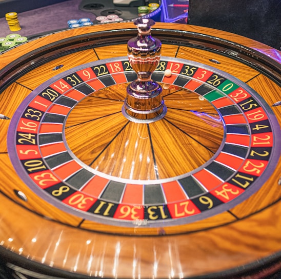 A roulette wheel with alternating red and black numbered pockets on a wooden platform. The wheel contains a small white ball resting in one of the pockets, indicating gameplay. Surrounding the wheel are stacks of colorful poker chips, suggesting a casino environment.