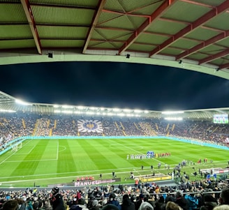 A large football stadium filled with spectators. The field is brightly lit, and two teams are positioned on the pitch. The stadium is covered, with a visible roof structure and stands filled with fans. Scoreboards are visible displaying game information.