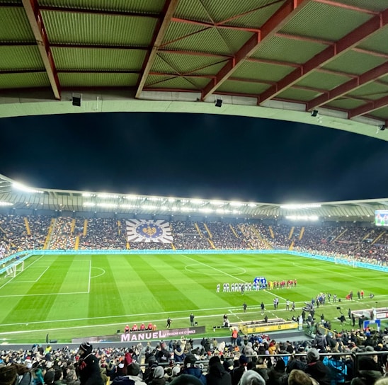 A large football stadium filled with spectators. The field is brightly lit, and two teams are positioned on the pitch. The stadium is covered, with a visible roof structure and stands filled with fans. Scoreboards are visible displaying game information.