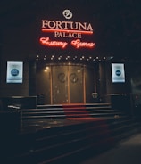 A neon-lit casino entrance with a red carpet leading up to large double doors. The sign reads 'Fortuna Palace Luxury Games' in bright red and white neon lights. The dimly lit atmosphere suggests a night-time setting. There are two digital displays on either side of the entrance showing promotional content.