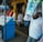 A street vendor stall displaying an array of lottery tickets clipped to a bright blue kiosk. A man in a red shirt leans on the counter, appearing thoughtful, while another man in a white shirt adjusts his hat and walks by. A variety of products hangs from strings above, and a colorful display fills the background with vibrant signs and packets. The scene suggests a busy, lively marketplace.