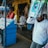 A street vendor stall displaying an array of lottery tickets clipped to a bright blue kiosk. A man in a red shirt leans on the counter, appearing thoughtful, while another man in a white shirt adjusts his hat and walks by. A variety of products hangs from strings above, and a colorful display fills the background with vibrant signs and packets. The scene suggests a busy, lively marketplace.